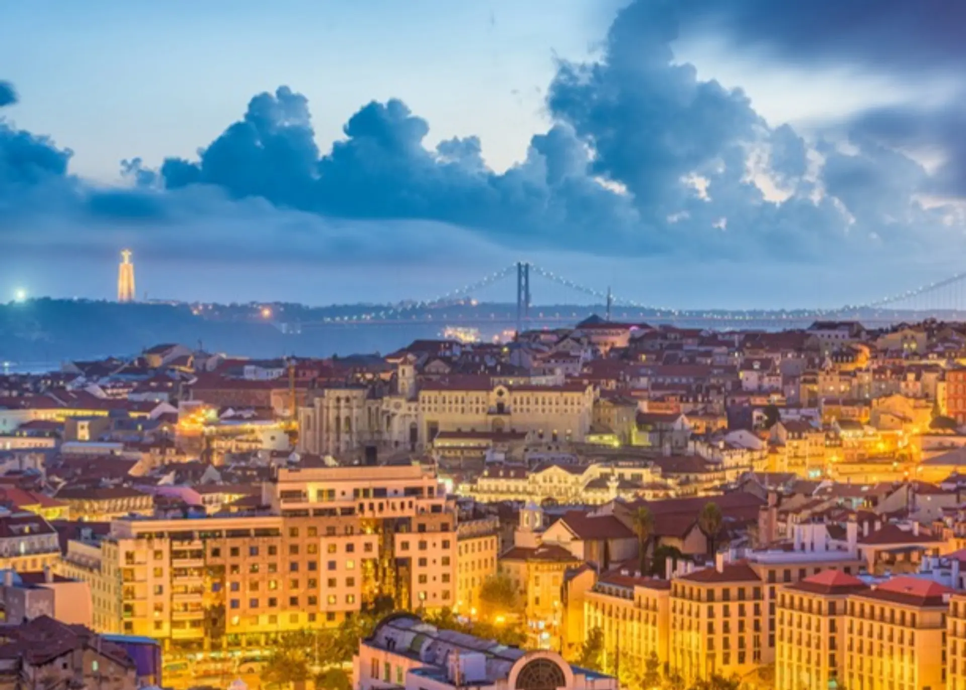 Lisbon's rooftops at dusk with the 25 de Abril Bridge and the Cristo Rei statue across the Tagus, for Sylvie di Giusto, international keynote speaker in Lisbon