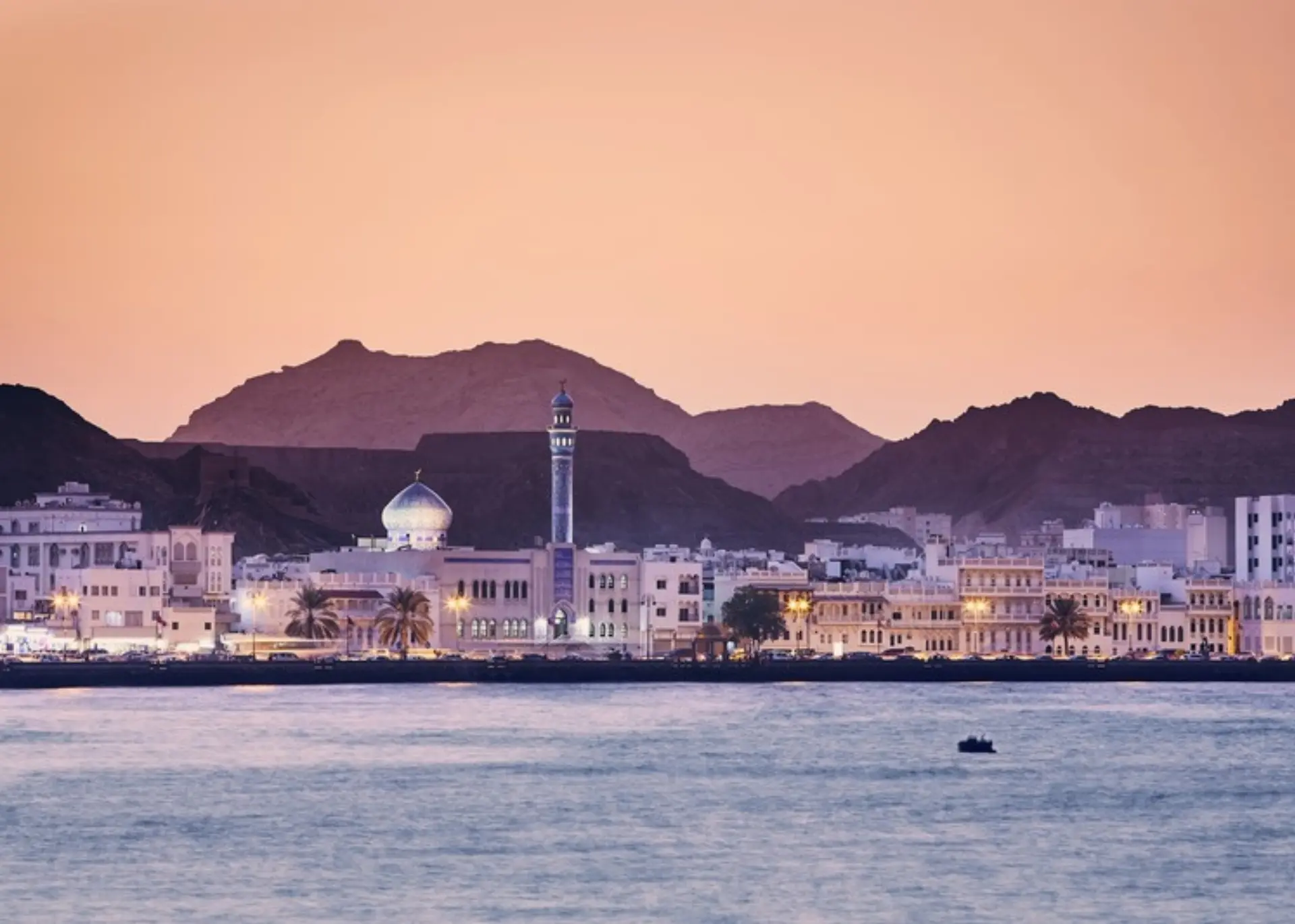 A mosque and the Mutrah waterfront in Muscat against the Hajar Mountains at dusk, for Sylvie di Giusto, international keynote speaker in Muscat